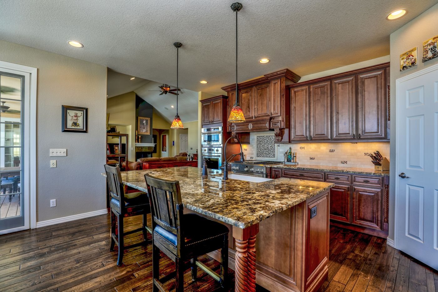 A kitchen with cherry cabinets, a granite island with two bar stools, a gas range and an opening through to a living room.