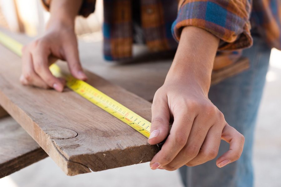 Two hands holding a tape measure along the edge of a rough sawn board.
