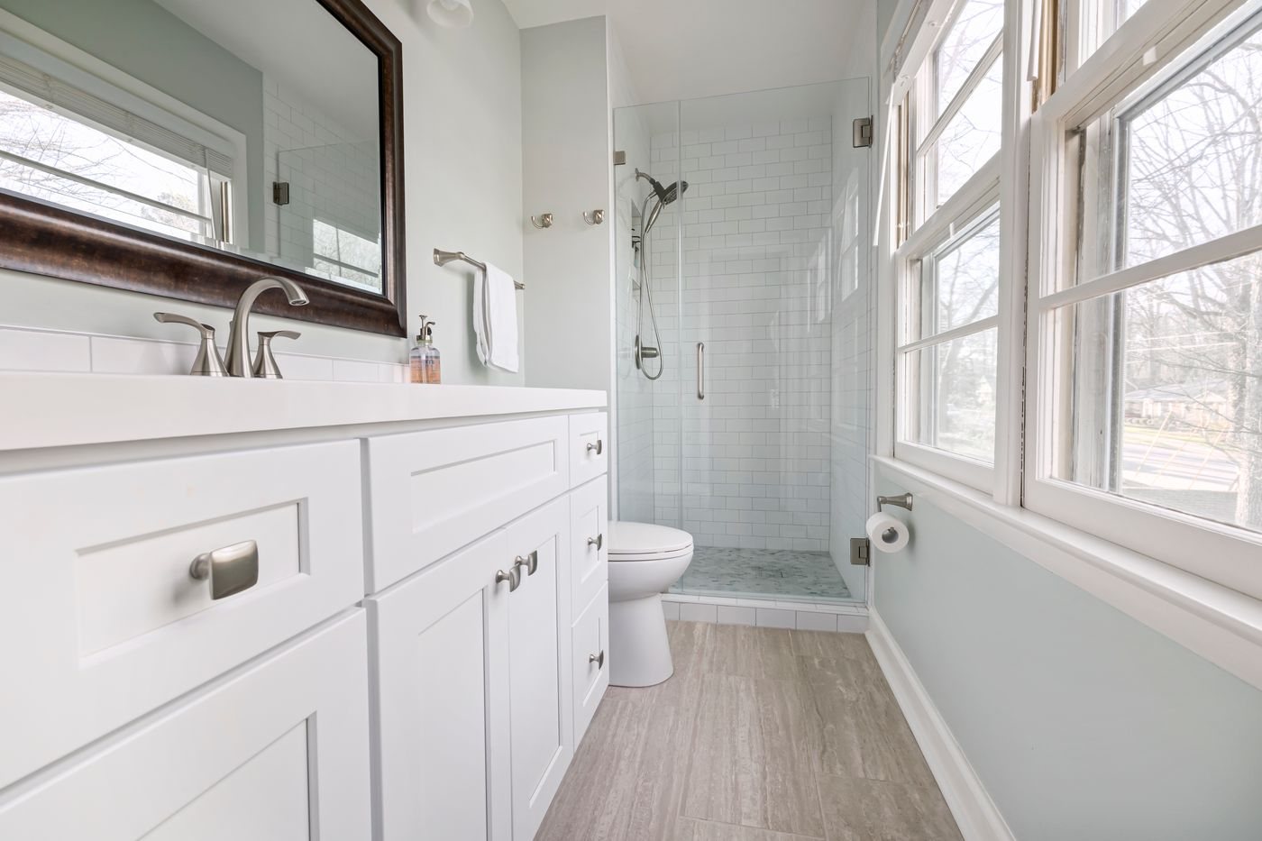 A bathroom with a white shaker vanity, quartz top, framed mirror and a subway tiled shower with a glass door, lit by a row of windows.