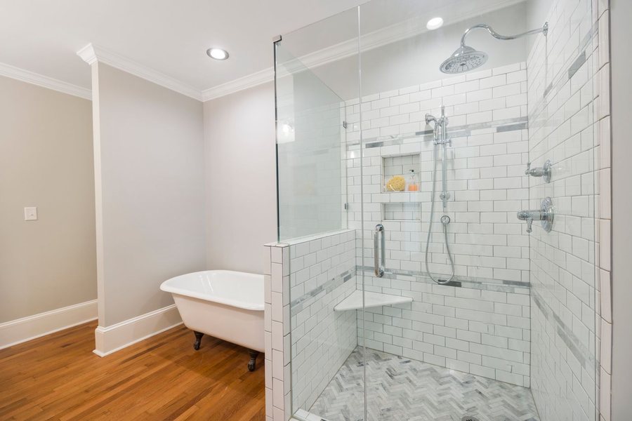 A large walk-in shower tiled in white subway tile with a marble herringbone floor, beside a freestanding tub on a hardwood floor.