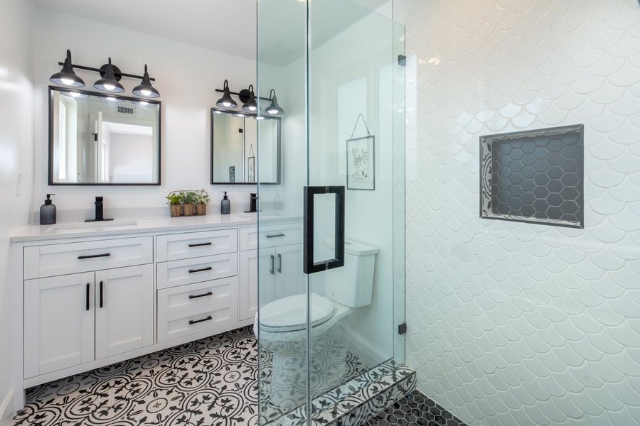 A bathroom with a white double vanity, black fixtures, two black framed mirrors, a patterned black and white floor and a white fish scale tile wall.