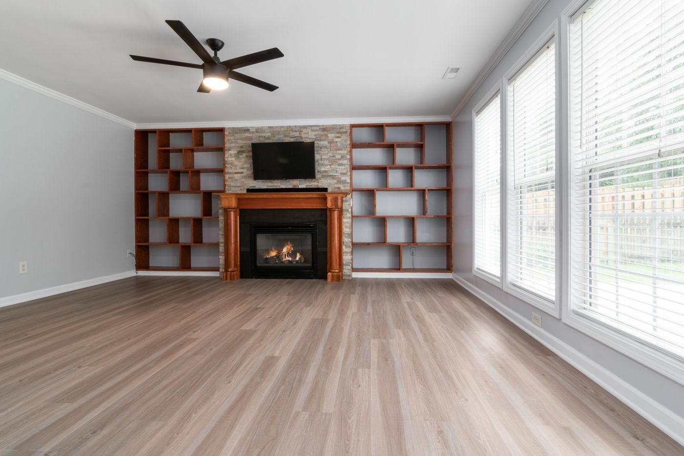 An empty family room with a stone faced fireplace, wooden built-in shelving on both sides, a ceiling fan and a bank of windows.
