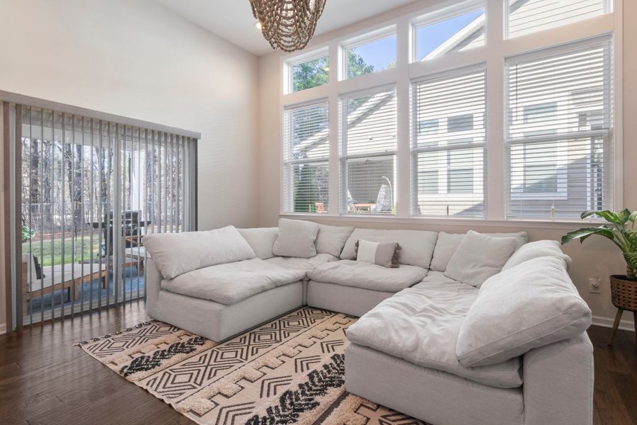 A bright sun room with a tall wall of windows, a pale sectional sofa on a patterned rug and a sliding door out to a deck.