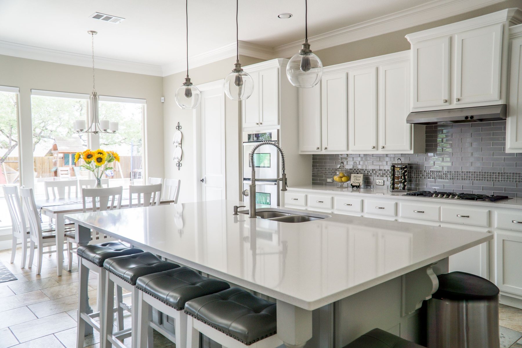 A finished kitchen with white cabinets, a large quartz island with four stools, glass pendant lights and a dining table beyond.