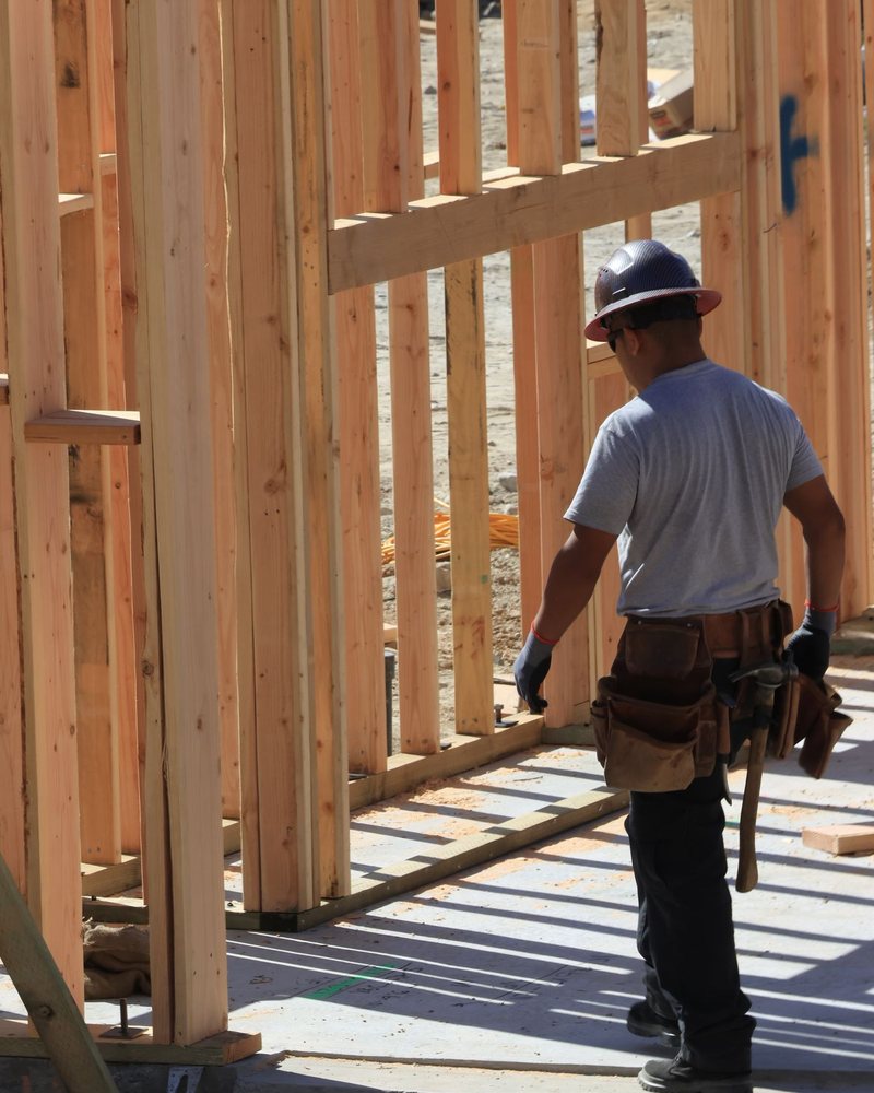 A carpenter in a hard hat and leather tool bags, seen from behind, standing in a newly framed stud wall on a concrete slab.