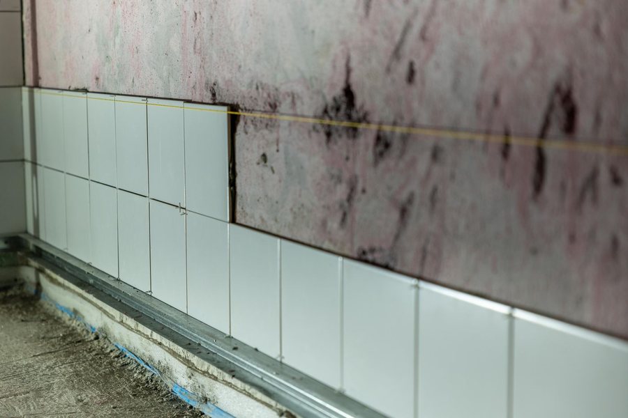 A wall part way through being tiled, with white tiles and spacers set against a pink waterproofing membrane above a bare concrete floor.