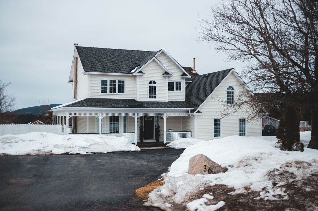 A white clapboard house with a wraparound porch behind a ploughed driveway and banked snow.