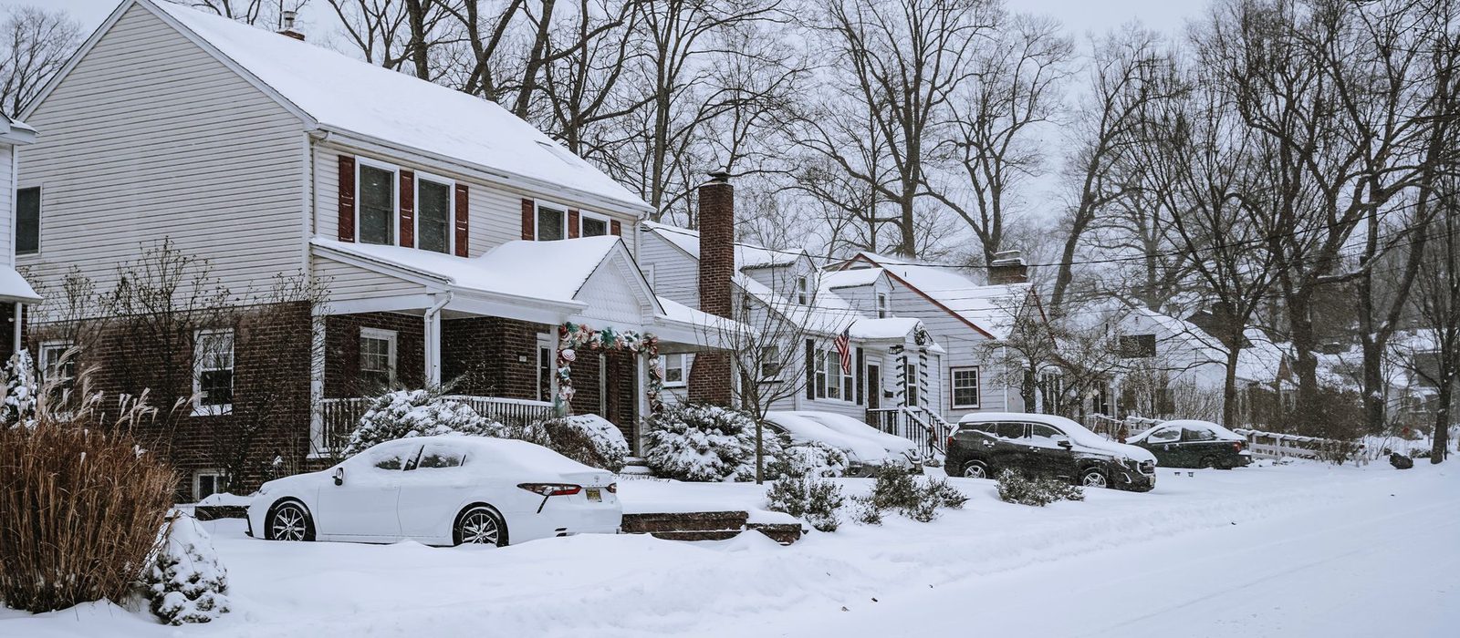 A suburban street after heavy snow, with cars buried at the kerb and snow on the roofs of clapboard houses.