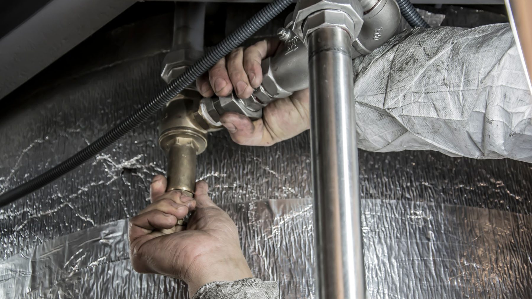 Two hands in white coveralls tightening a brass and steel pipe fitting behind a foil insulated wall.