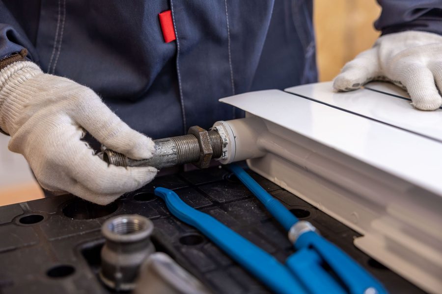 Gloved hands guiding a threaded steel pipe connection into a white panel radiator, with two pipe wrenches on the bench.