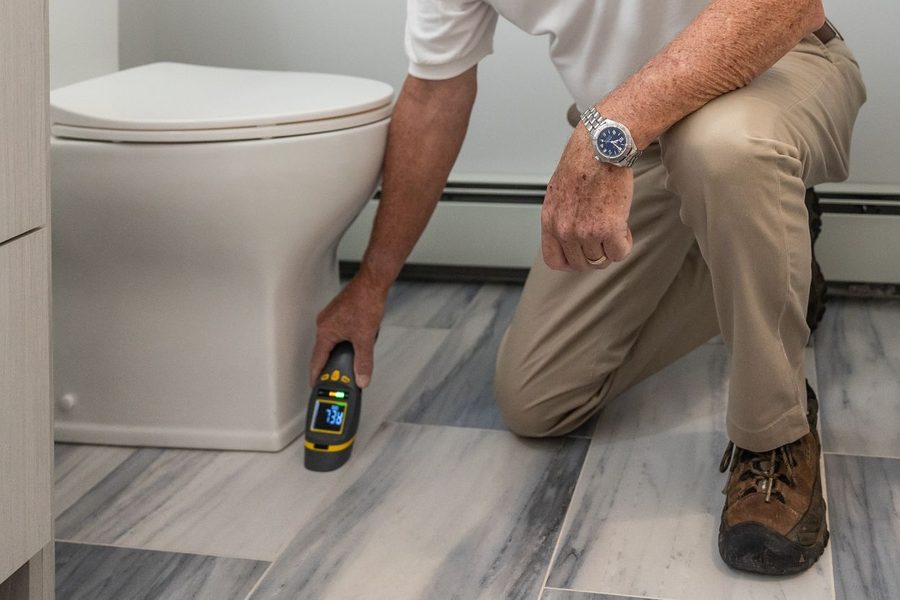 A kneeling worker holding a digital moisture meter reading 73.6 against tile at the base of a toilet.