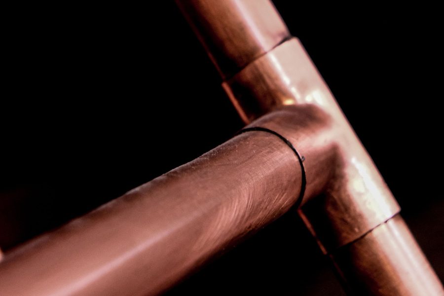 A soldered copper tee joining three lengths of copper pipe, lit against a black background.