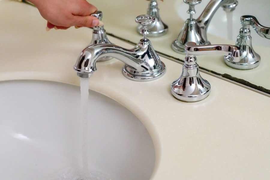 A hand turning the handle of a chrome widespread faucet while water runs into a cream colored basin.