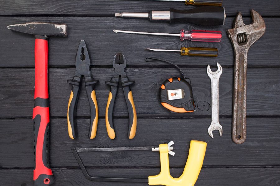 Hand tools laid out on dark painted boards: a hammer, pliers, screwdrivers, a tape measure, a hacksaw and an adjustable wrench.