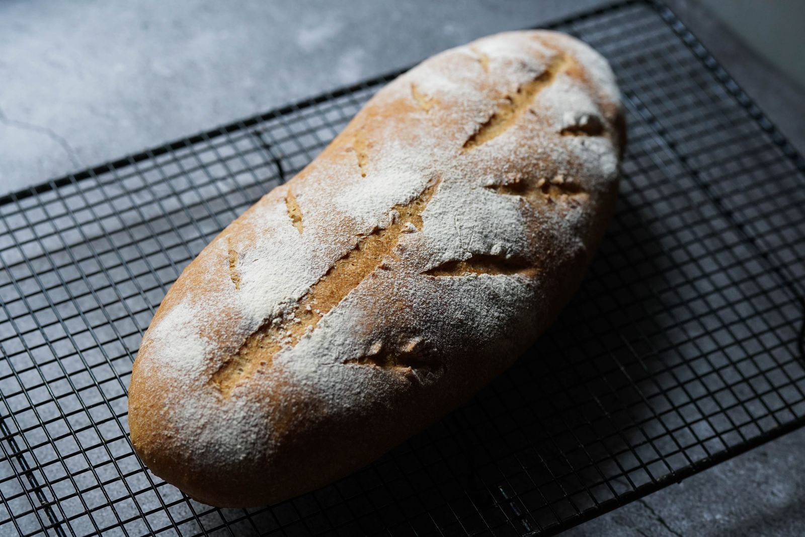 A single flour dusted loaf with slashed scoring, resting on a black wire cooling rack.