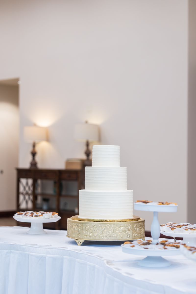 A three tier cake with plain white buttercream on a gold stand, with plates of pastries on either side.