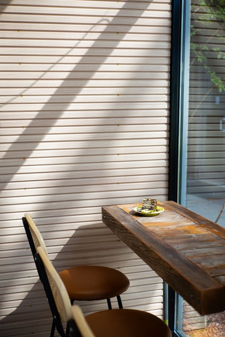 A reclaimed wood ledge table with two stools in a sunlit corner, a slice of layered cake on a plate at one end.