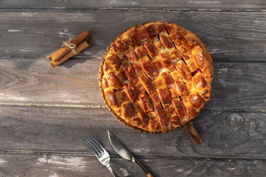 A whole lattice topped apple pie on a round wooden board with cinnamon sticks and cutlery on gray planks.