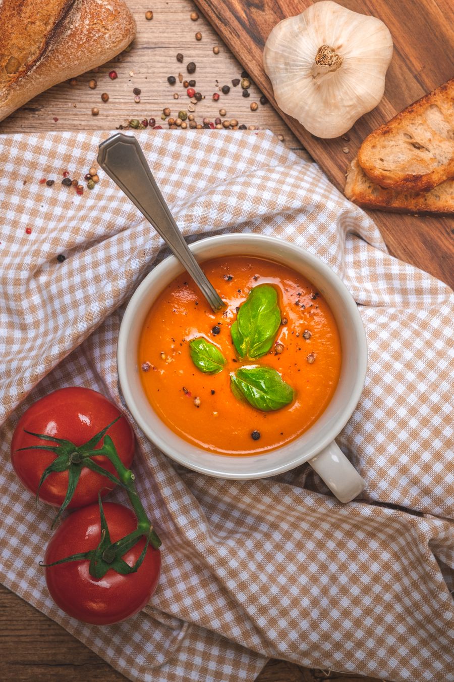 A white cup of tomato soup topped with basil leaves, on a checked cloth with vine tomatoes and toasted bread.