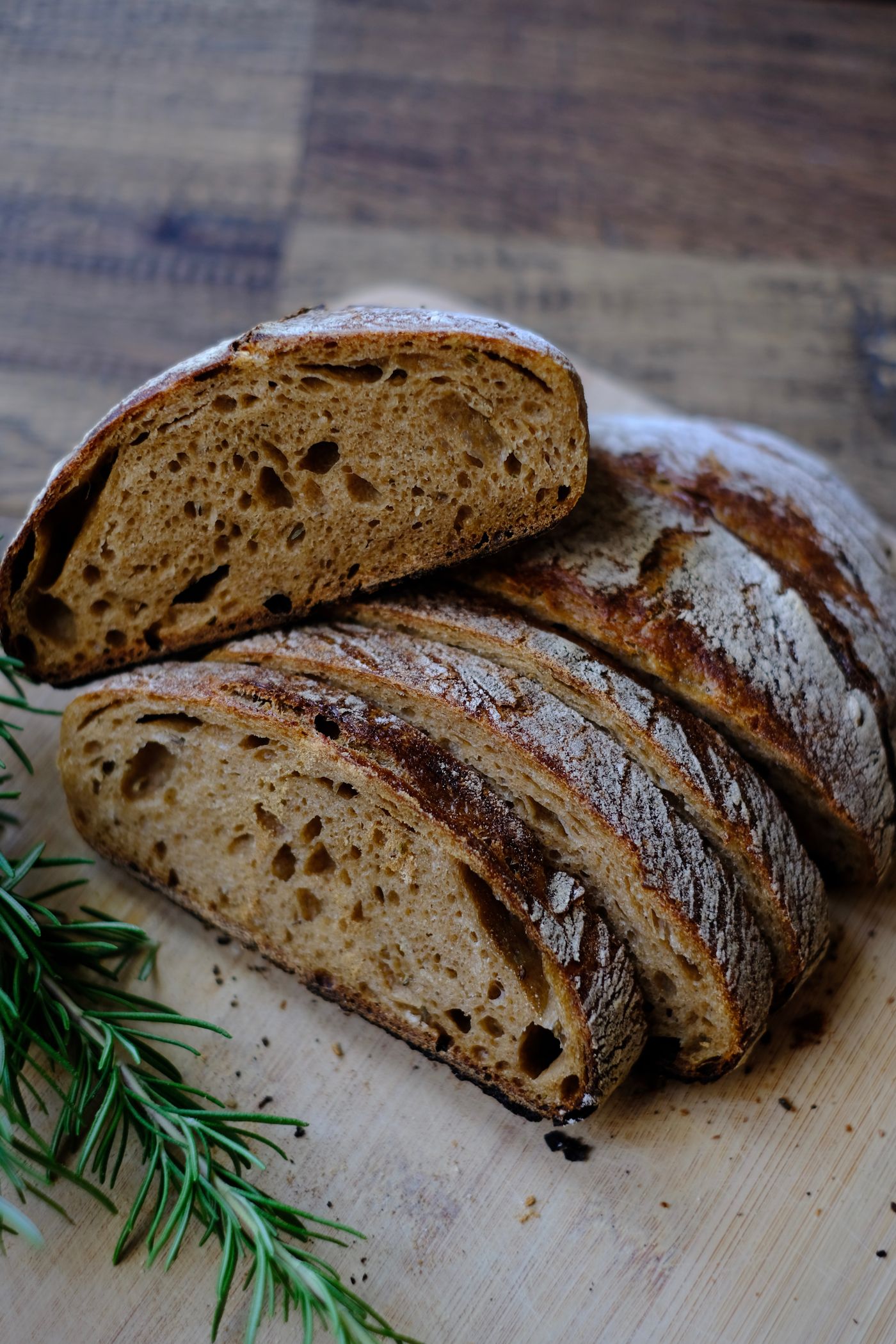 A round country loaf cut open on a wooden board, showing an open crumb, with a sprig of rosemary beside it.