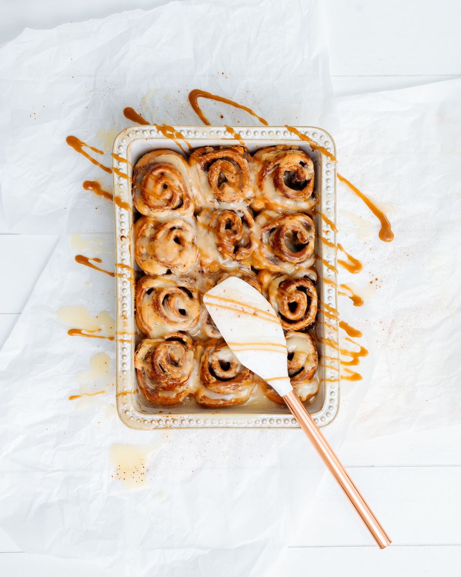 A tray of glazed cinnamon rolls in a baking dish on white parchment, with caramel drizzled around it.