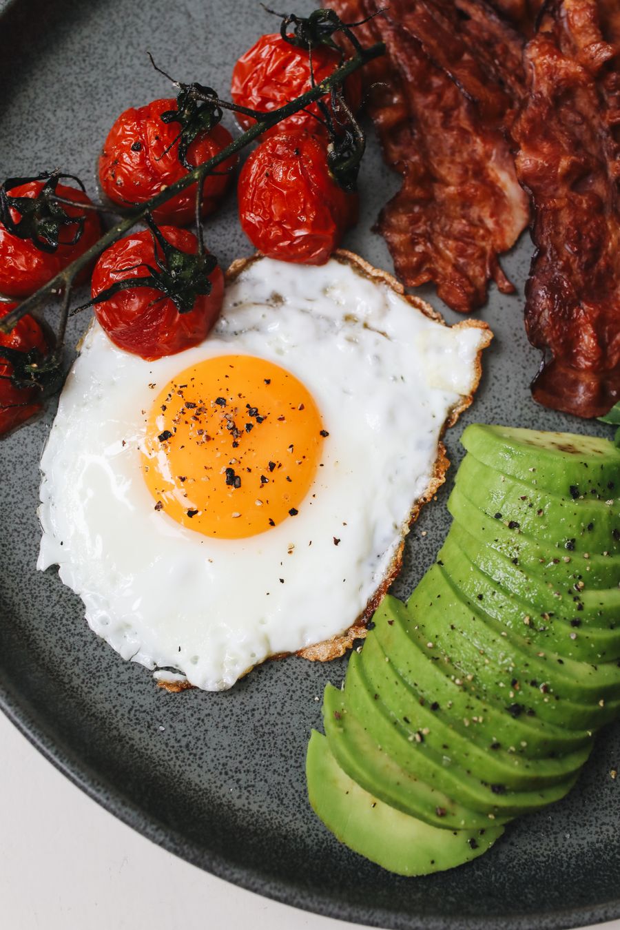 A fried egg with a bright yolk, strips of bacon, roasted vine tomatoes and fanned avocado slices on a dark speckled plate.