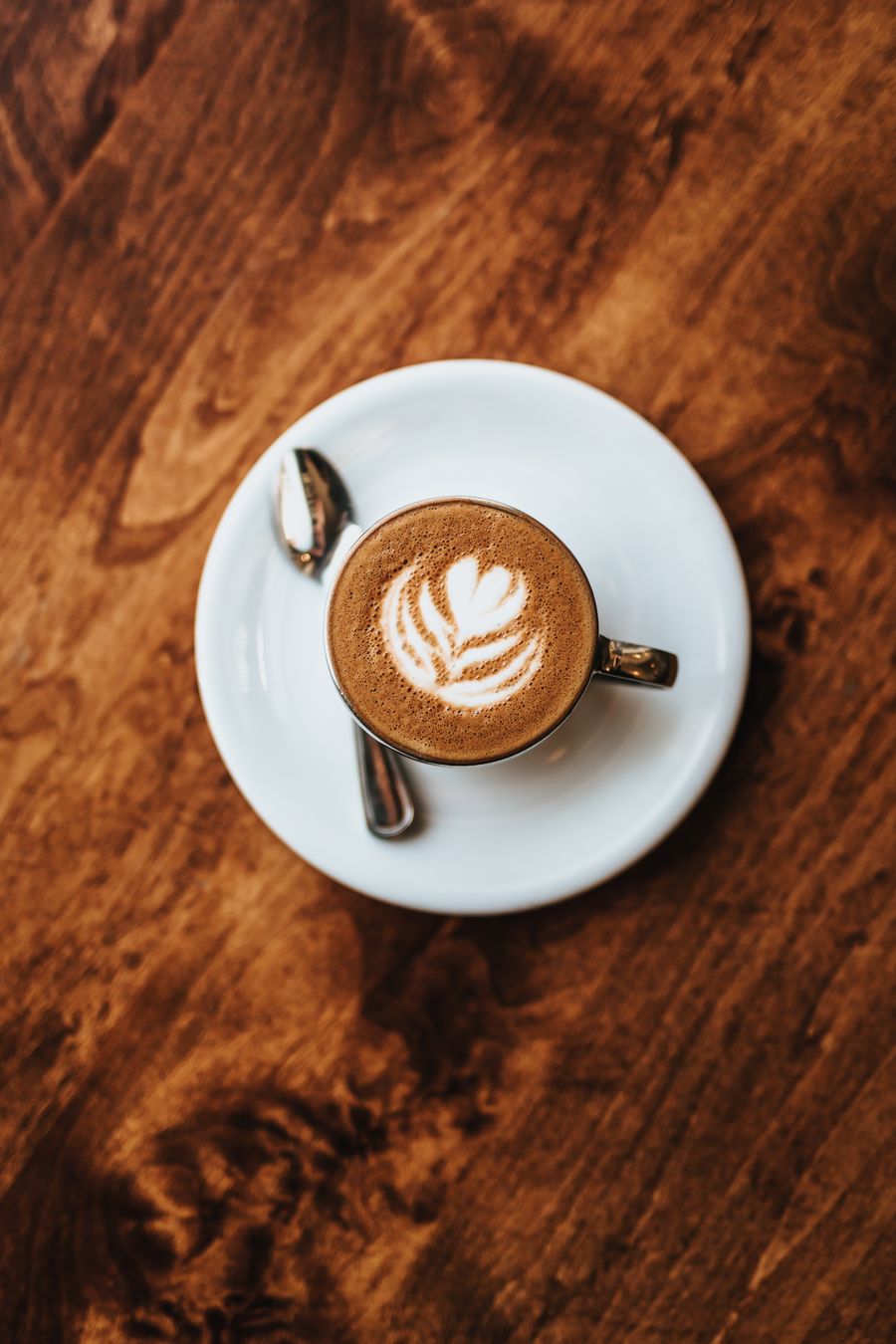 A cappuccino with a leaf poured in the foam, on a white saucer set on a dark polished wood table.