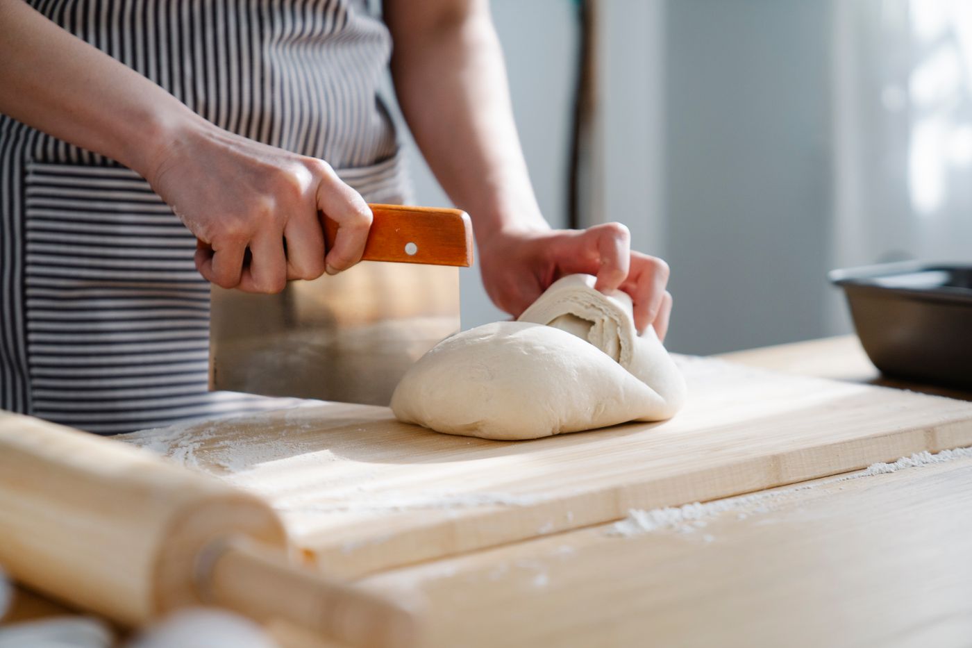 Two hands dividing a round of bread dough with a metal bench scraper on a floured wooden board.