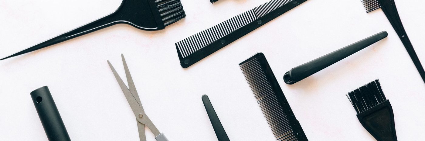 Black cutting combs, shears, a tail comb and tint brushes laid out in a row on a pale surface.
