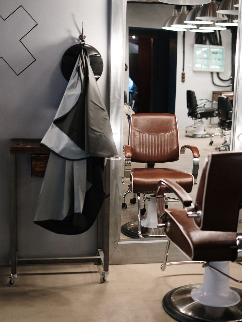 Two brown leather styling chairs in front of a tall mirror with a cape hanging beside them.