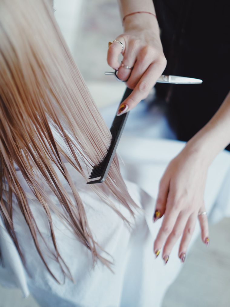 A comb drawn through long wet blonde hair over a white cape, shears held ready.