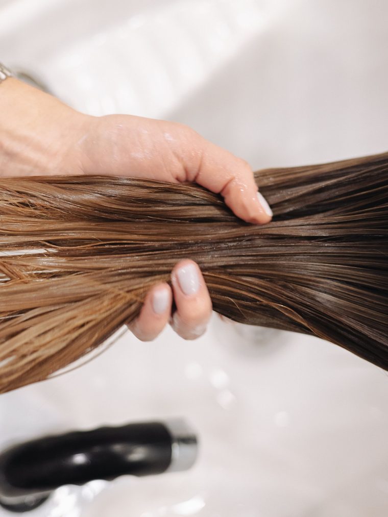 Hands lifting a length of wet brown hair over a white basin during a rinse.