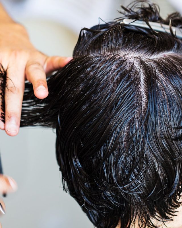A stylist lifting a section from the back of a head of wet dark hair, comb held against it.
