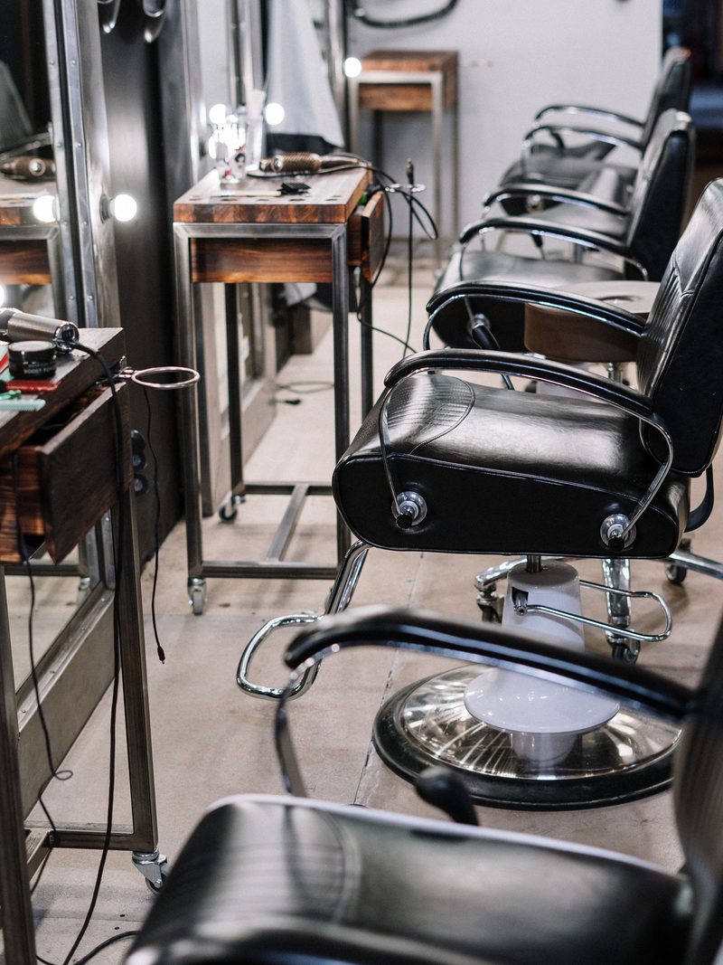A row of black styling chairs facing wooden stations and lit mirrors on a concrete floor.